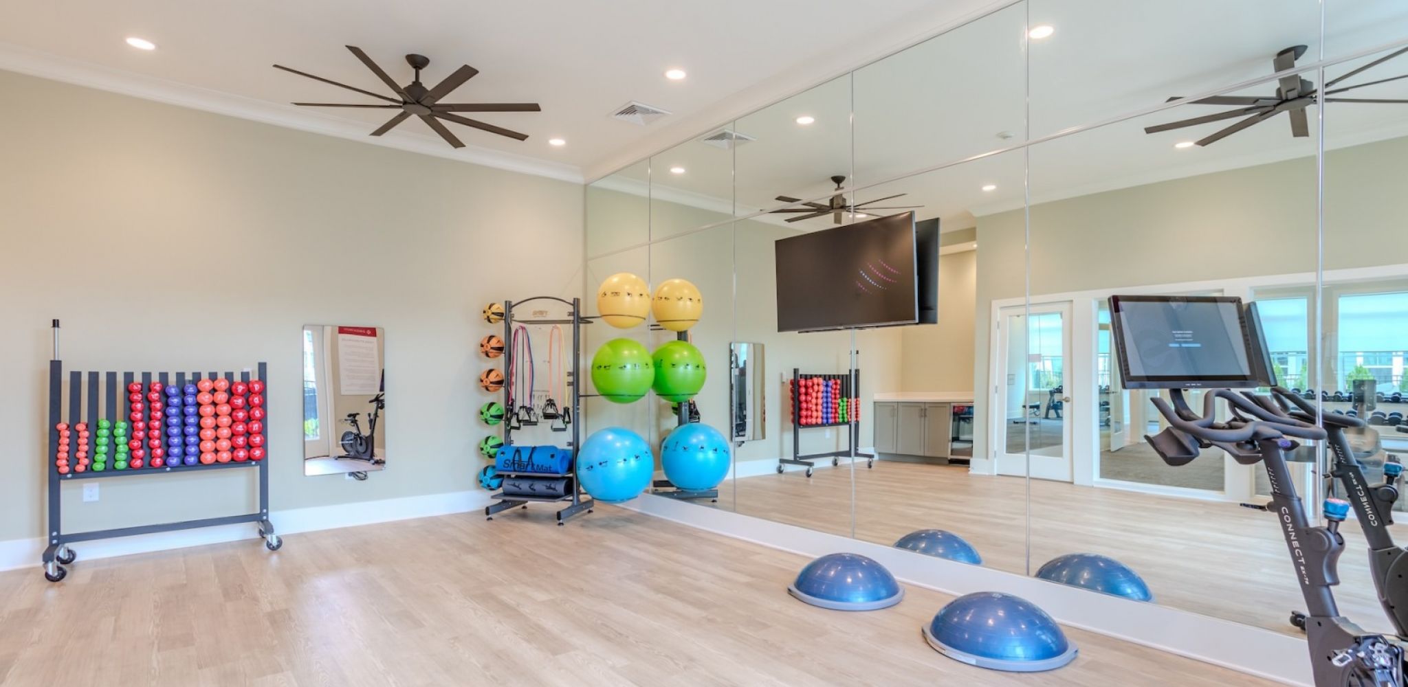 Hawthorne at New Bern bright fitness room with exercise balls, weights, mirrors, a bike, TVs, and wooden floors.