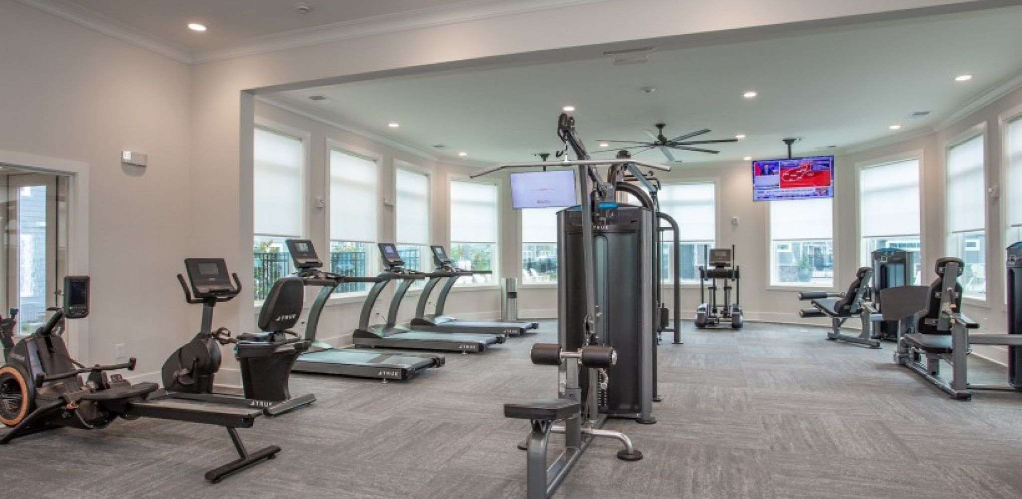 Hawthorne at New Bern modern gym with cardio machines, strength equipment, large windows, and a TV on the wall.