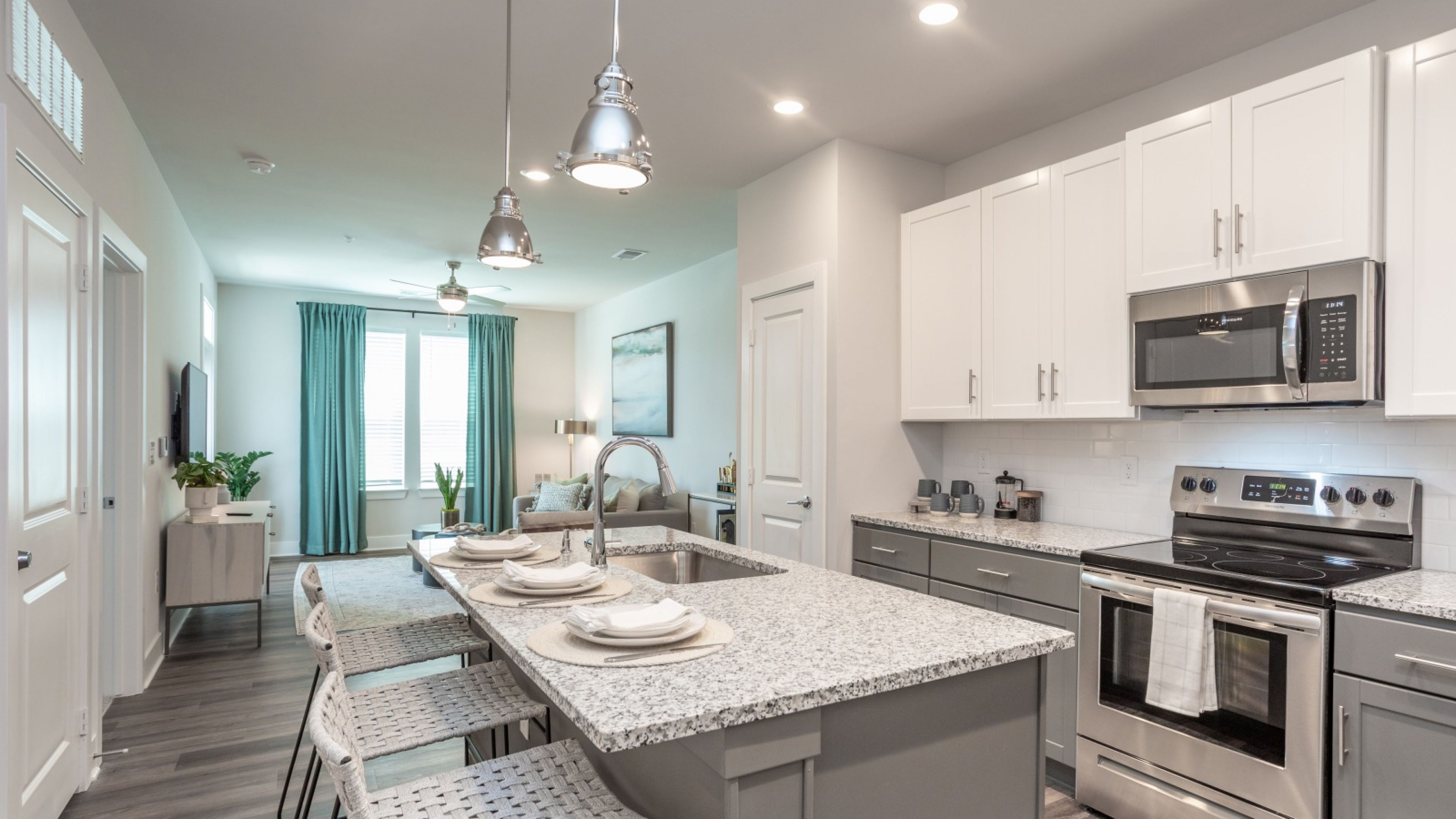 Hawthorne at New Bern modern kitchen with granite island, white cabinets, bar stools, and view into a bright living room with blue curtains.