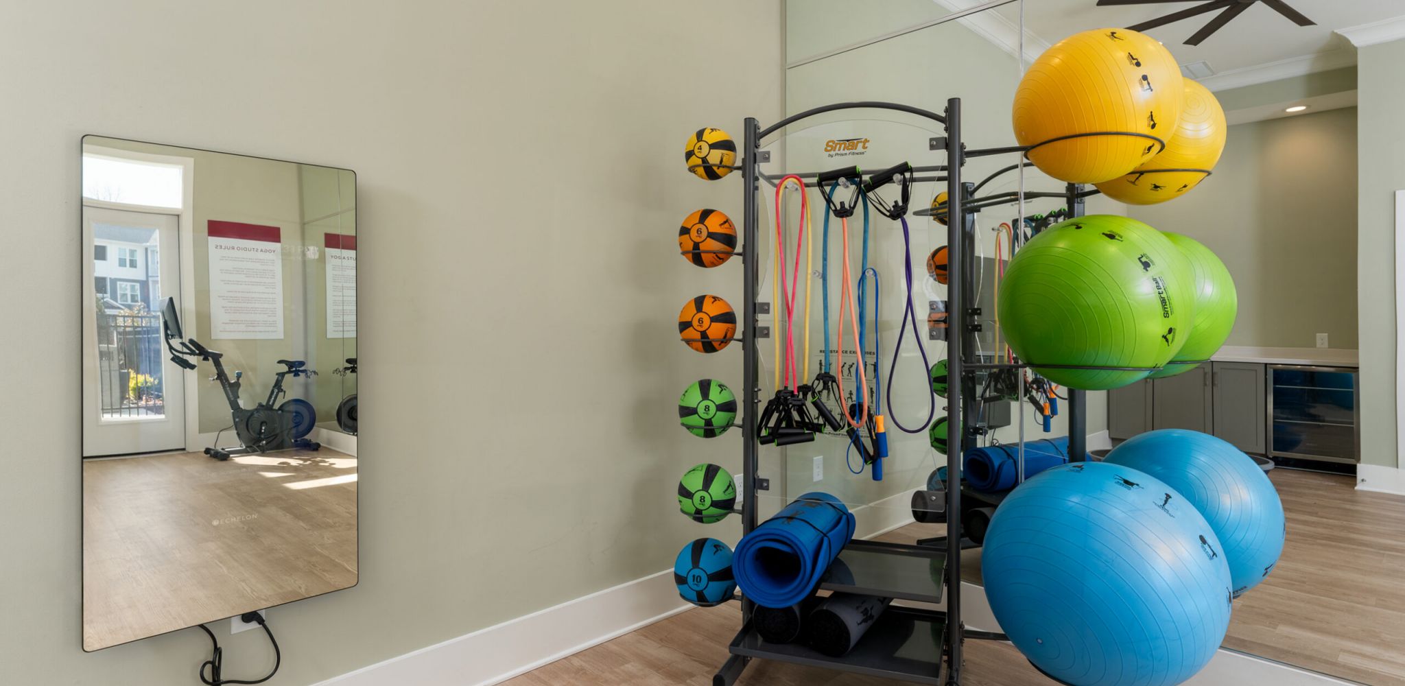 Hawthorne at New Bern bright fitness room with exercise balls, weights, mirrors, a bike, TVs, and wooden floors.