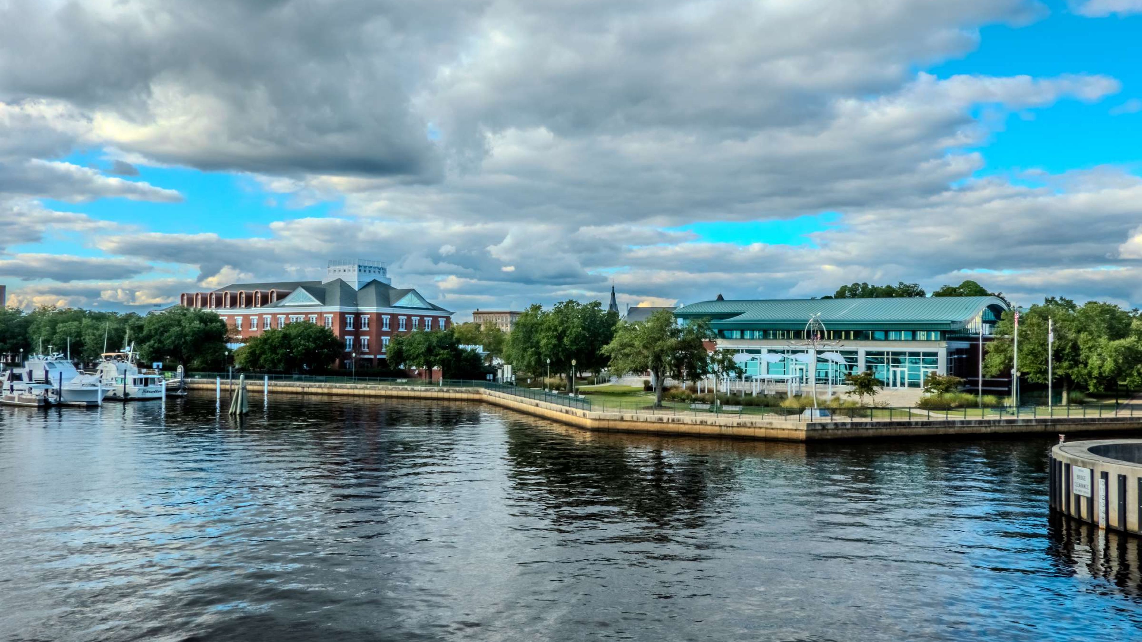Hawthorne at New Bern waterfront with boats, modern and brick buildings, green trees, and a partly cloudy blue sky.